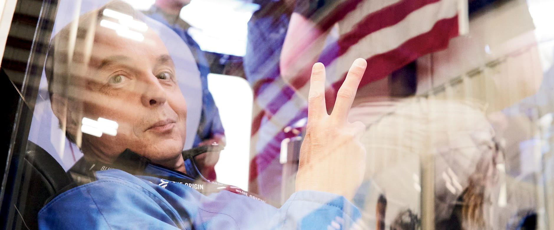 A crew member of Blue Origin's space exploration program giving a peace sign through a window