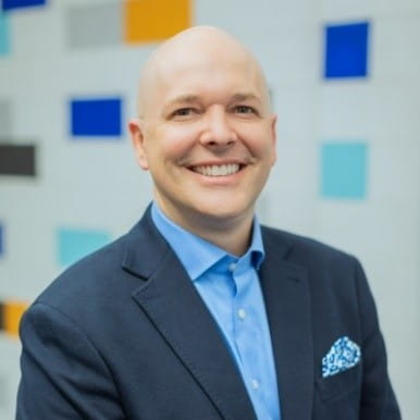 A bald white man in a dark grey suit and light blue button down grins at the camera.