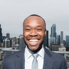 Ezeibe, a Black man in a suit and tie, smiles widely at the camera against the city skyline background.