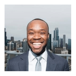 Ezeibe, a Black man in a suit and tie, smiles widely at the camera against the city skyline background.