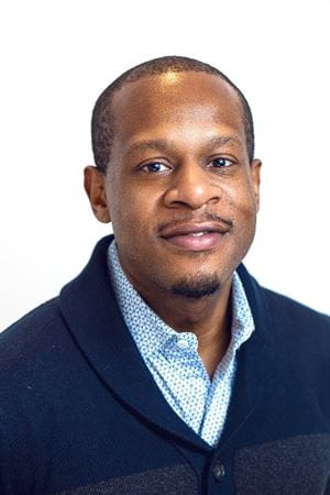 Fritz, a black man in a patterned blue button down and navy blue sweater, smiles softly at the camera.
