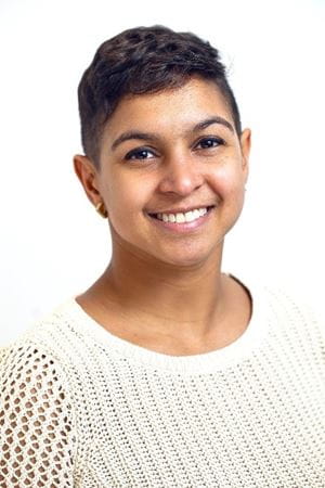 Sydney, a Black woman with short hair, smiles brightly at the camera. She wears a cream knit sweater and small earrings.