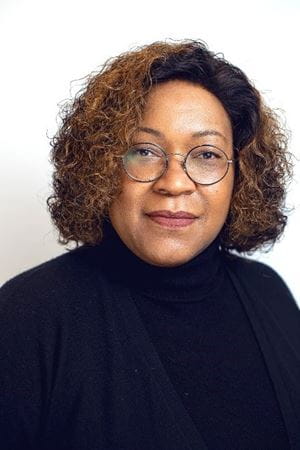Tewana, a Black woman in a black turtleneck and round glasses, looks softly at the camera.