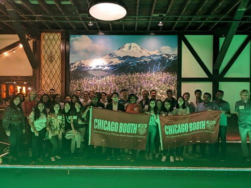 A group of 36 alumni smile at the camera, holding two Chicago Booth flags, while standing in front of a large painting of a snowcapped mountain and a field of wildflowers.