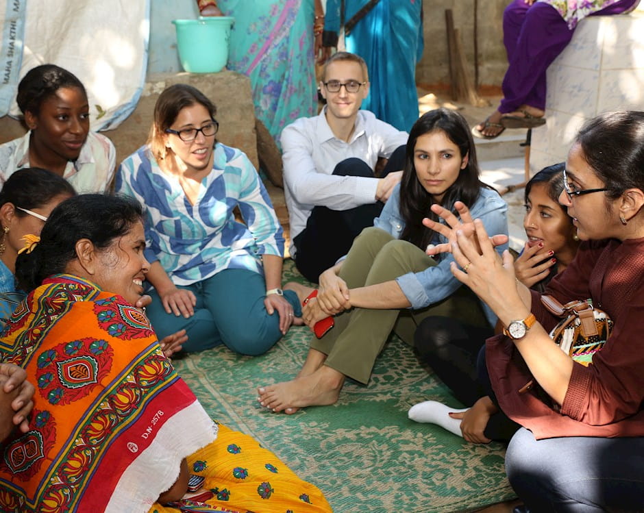 Students sit on a patterned rug in a circle, listening to women wearing Indian saris