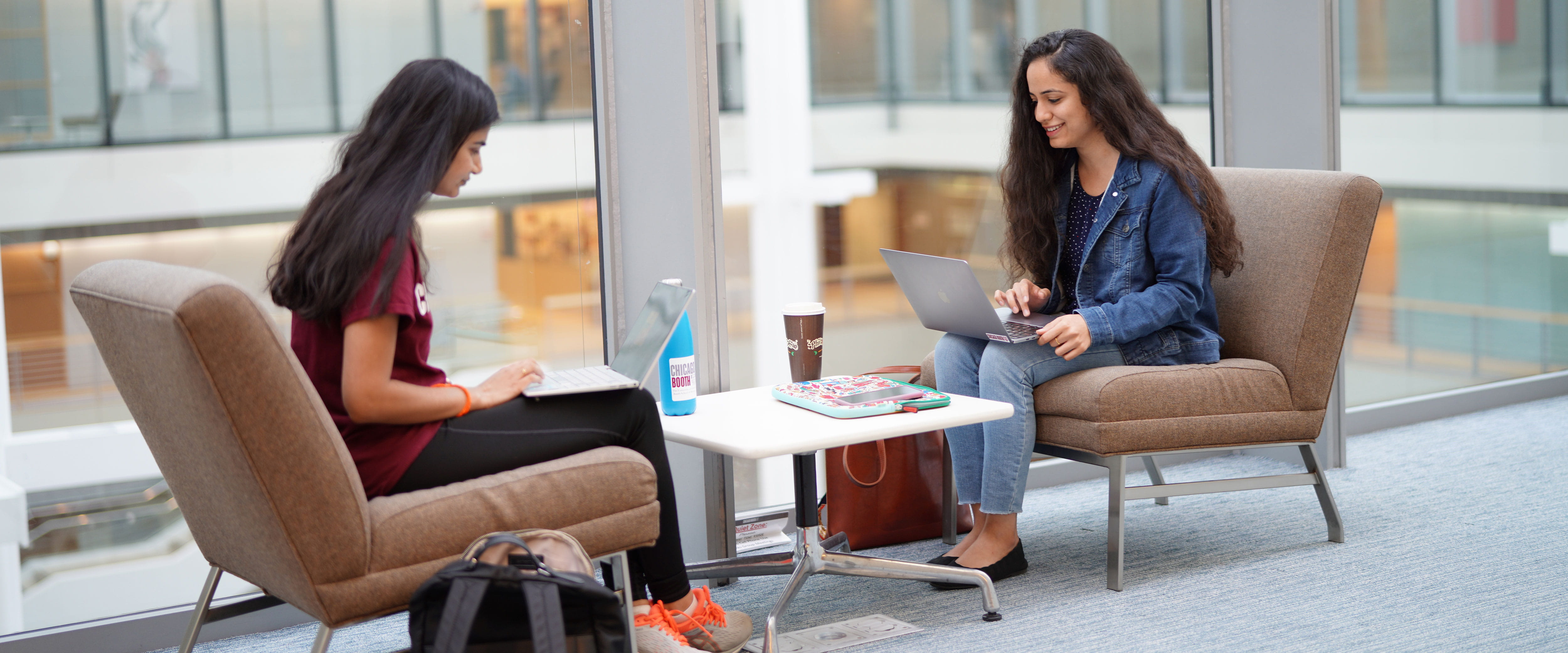 Two women sitting in brown chairs directly across from one another on their laptops 