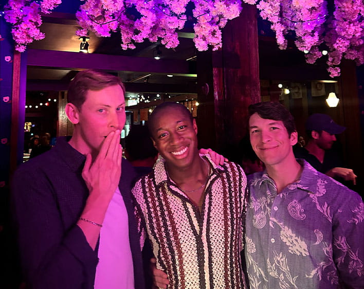Deasee and two other Boothies posing with one Boothies hand covering their mouth. Background has pink neon lighting and flowers hanging down from ceiling.
