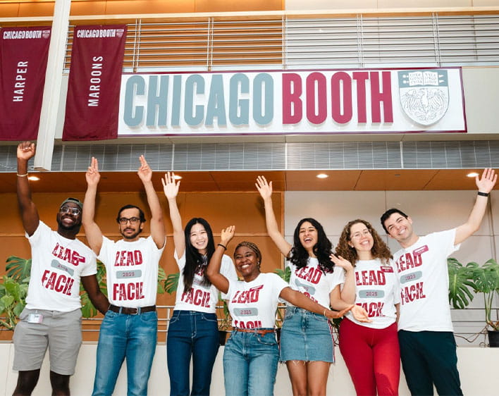Ope and six of his fellow Boothies wearing Lead Facil 2025 t-shirts while smiling together with Chicago Booth signage in background