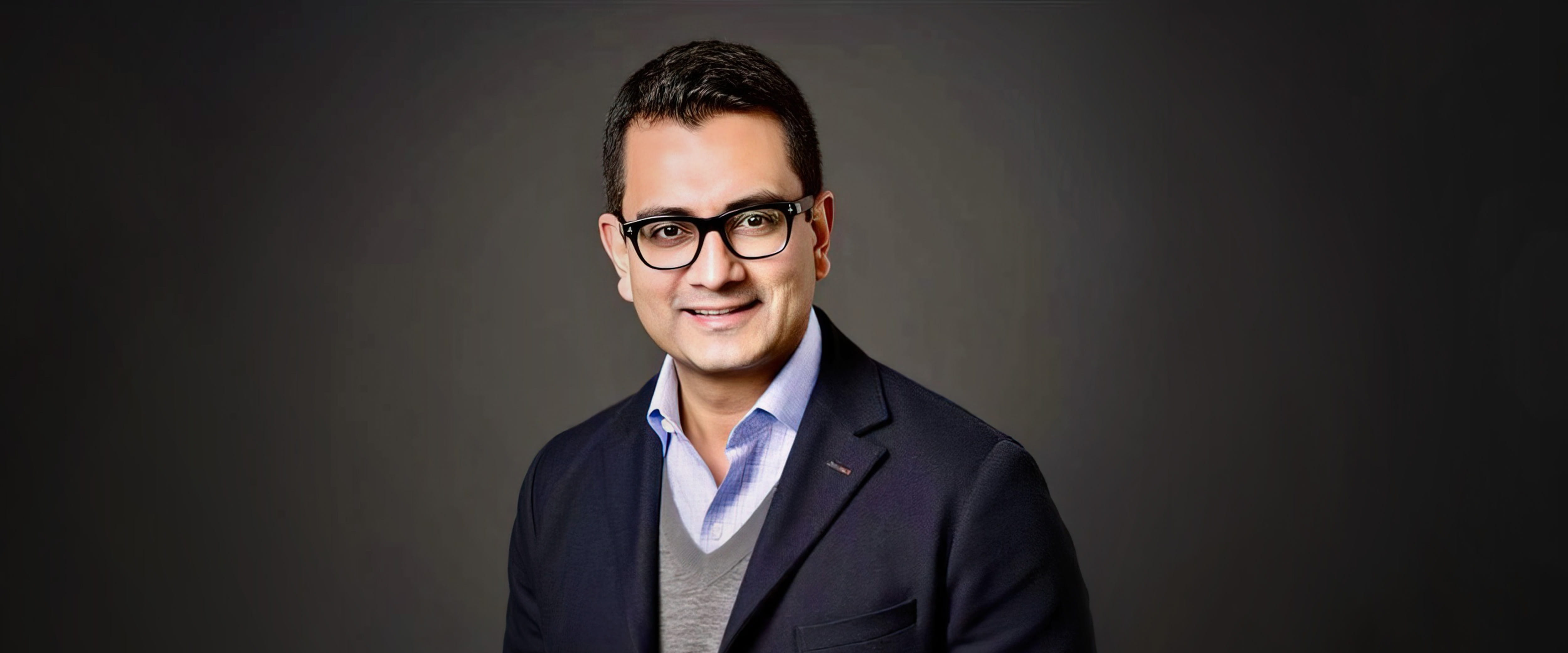 Chicago Booth alumnus JG Chirapurath wearing a blazer and smiling against a dark background.