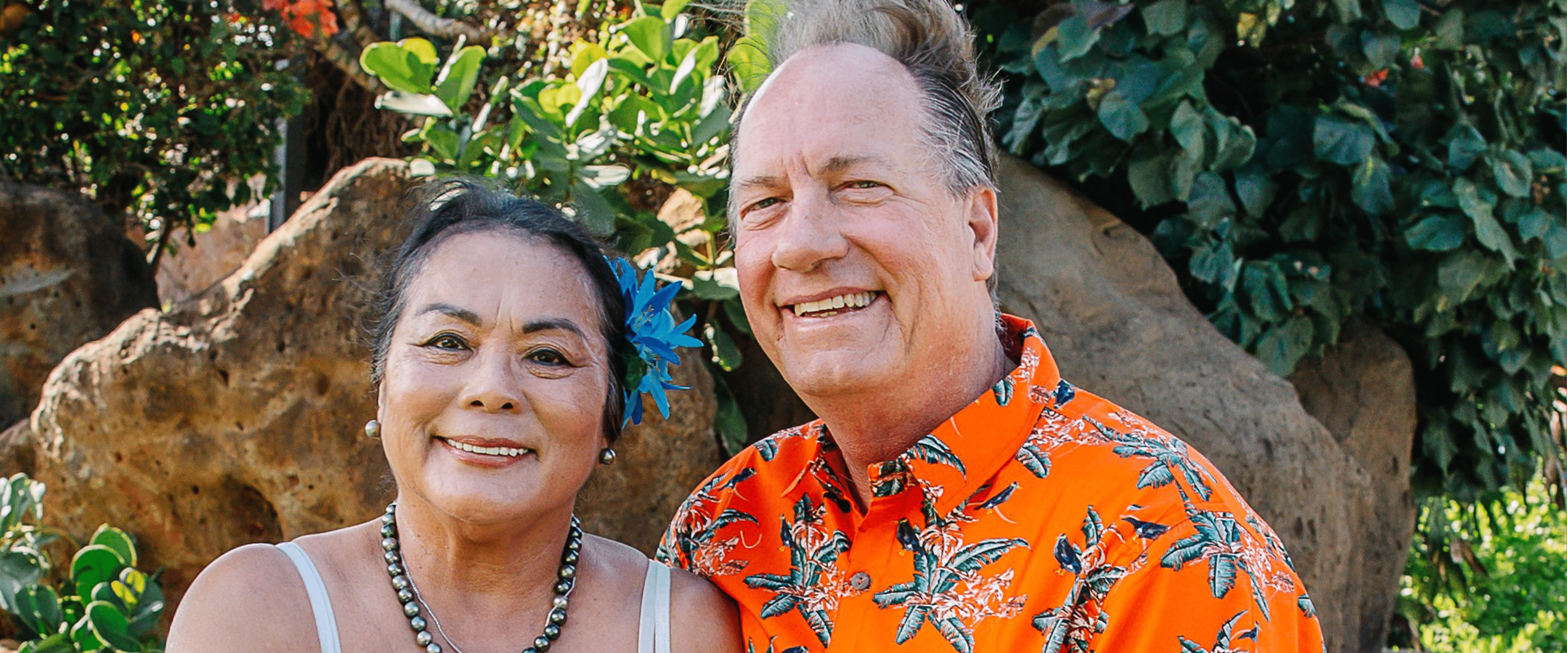Alumnus Larry Popelka and his wife in beachwear standing in front of a tropical tree.