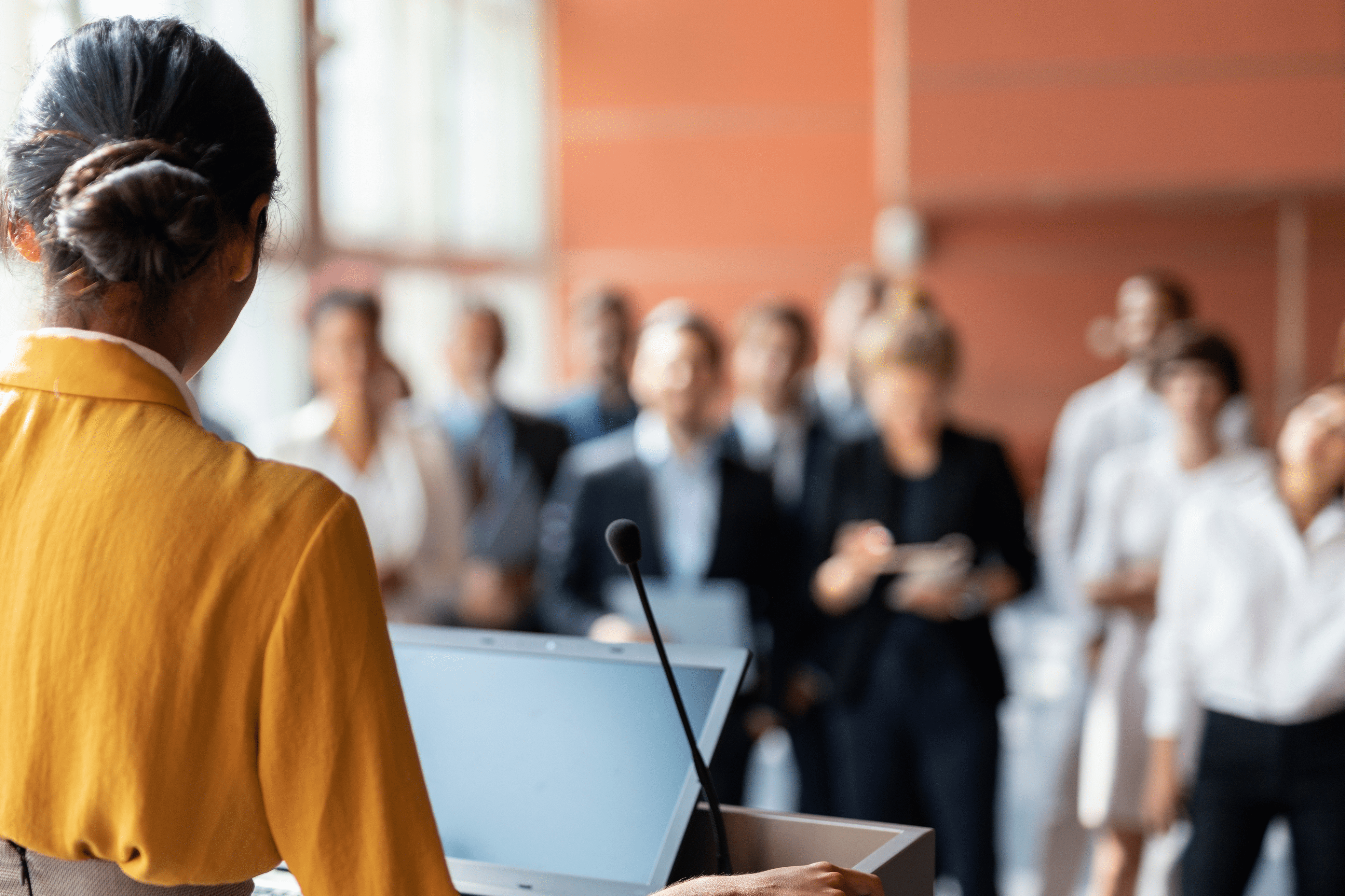 Woman with brown hair and yellow suit jacket speaks at a podium in front of an audience