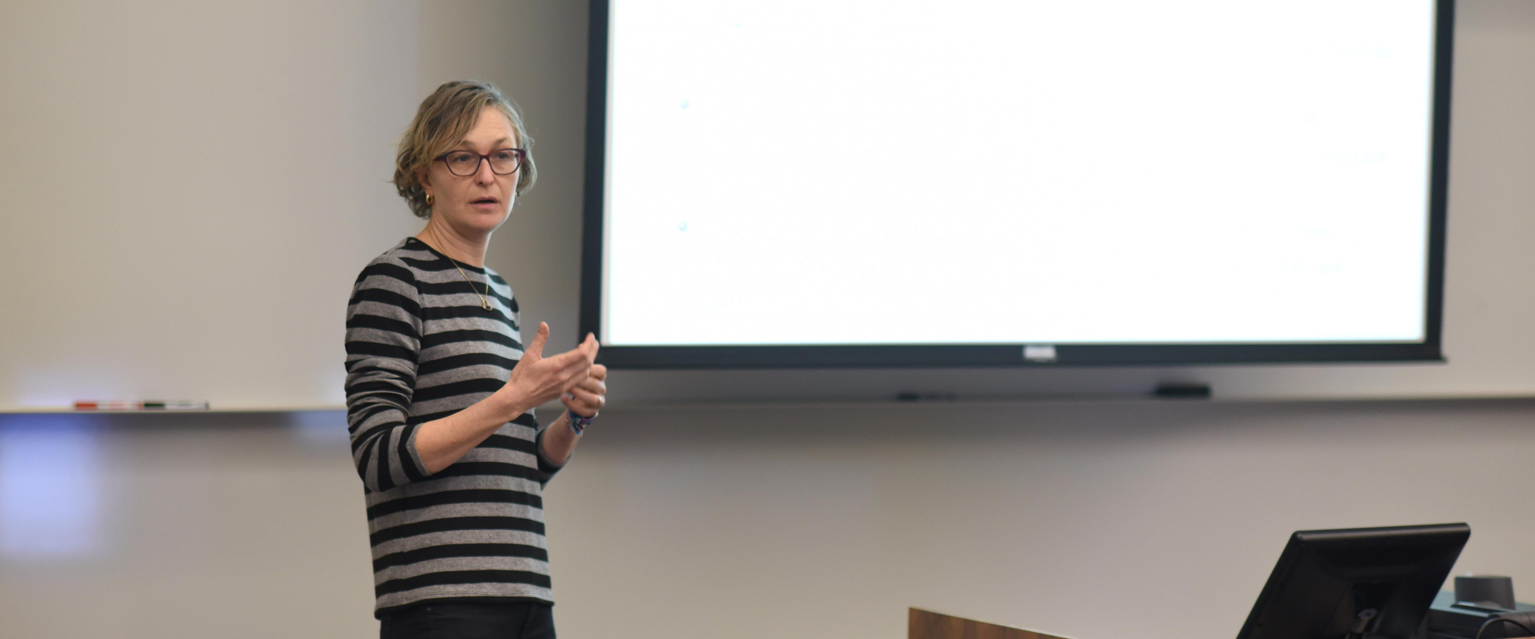 Marianne Bertrand speaking at the front of a classroom in frontof a projector screen