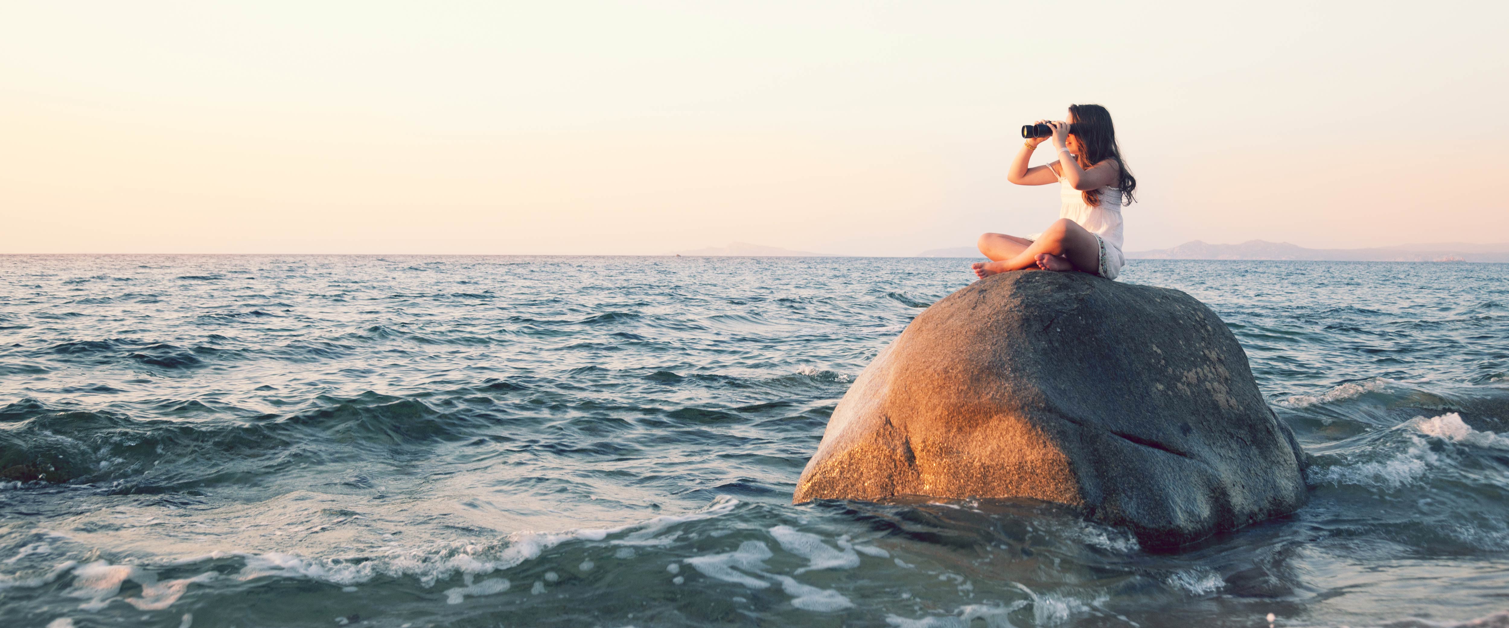 A young person looks out at the water with binoculars from the top of a boulder