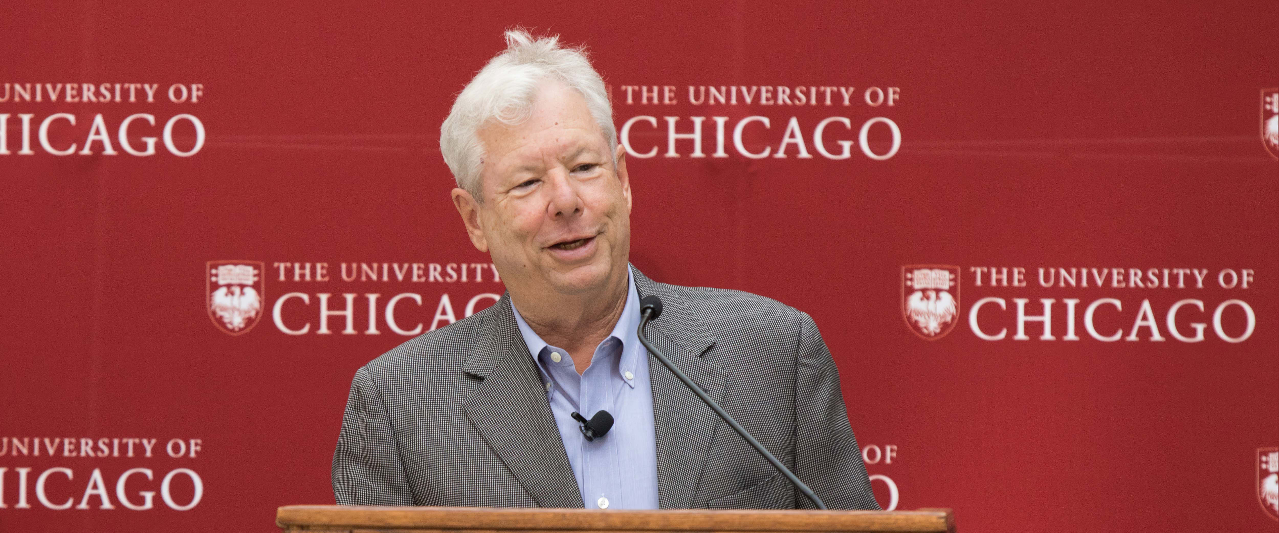  Richard H Thaler speaking at a podium in front of a maroon, university-branded background in 2017