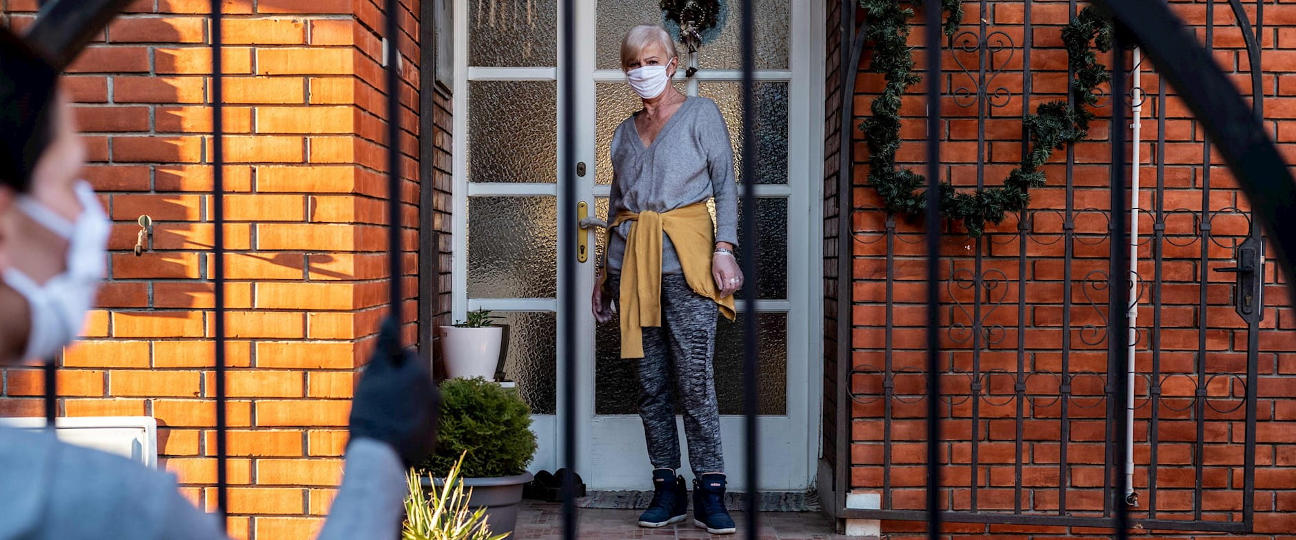 A masked silver-haired woman on her porch waits for a grocery delivery from a masked volunteer