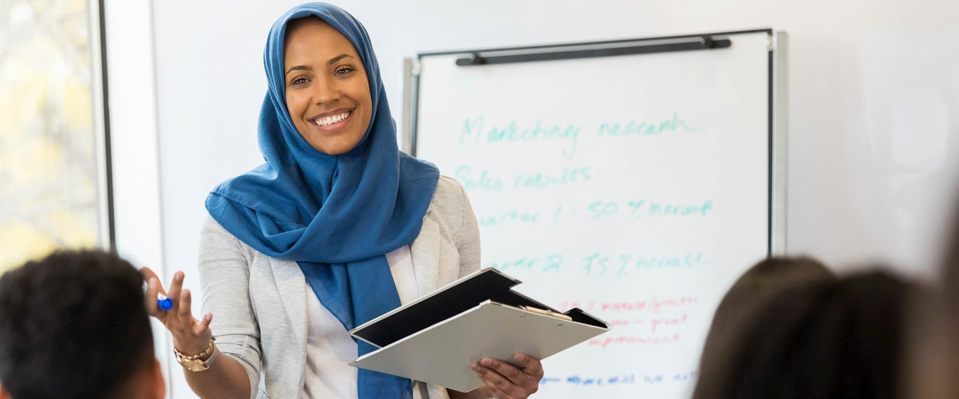 Smiling businesswoman in a blue hijab holding a clipboard in front of a dry erase board; she's talking with seated colleagues
