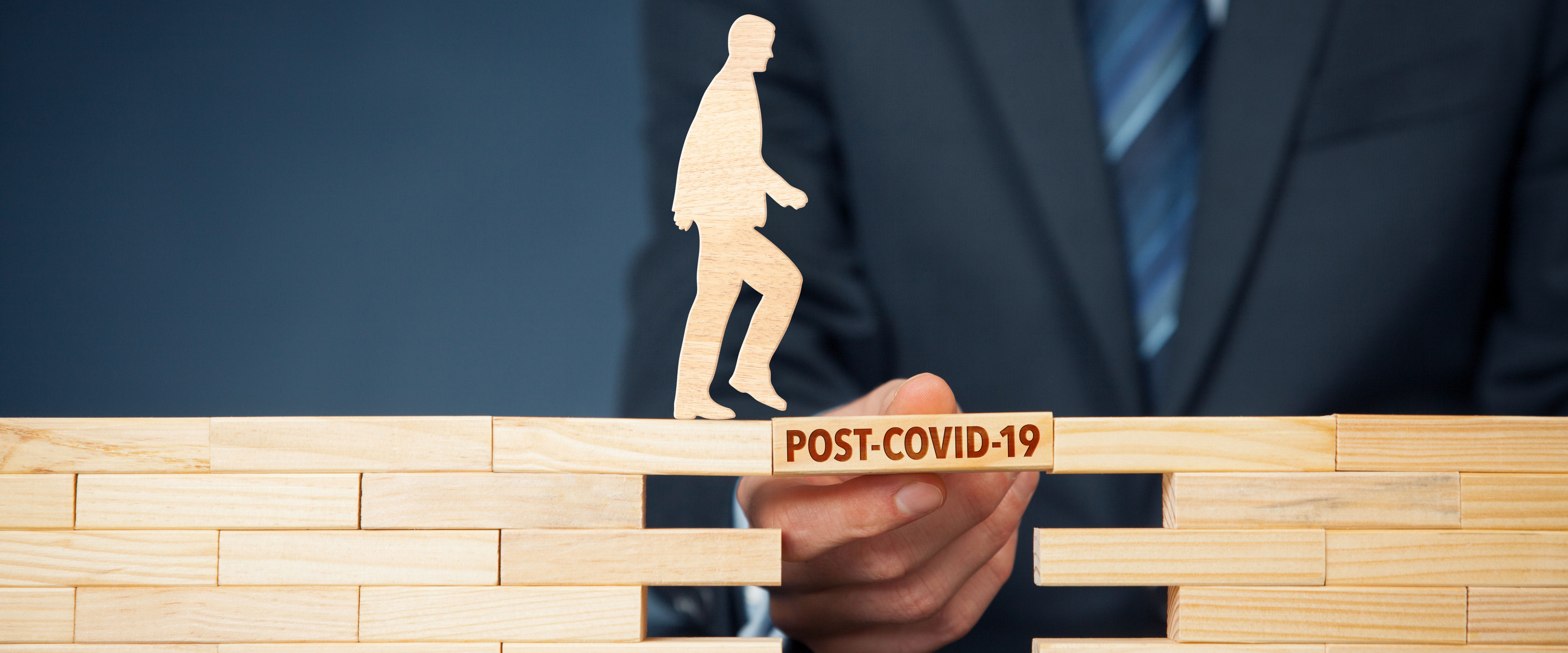 A businessman using his hand to build a mini wooden bridge across a gap - the bride is labeled "Post COVID-19" and a small wooden person is about to cross