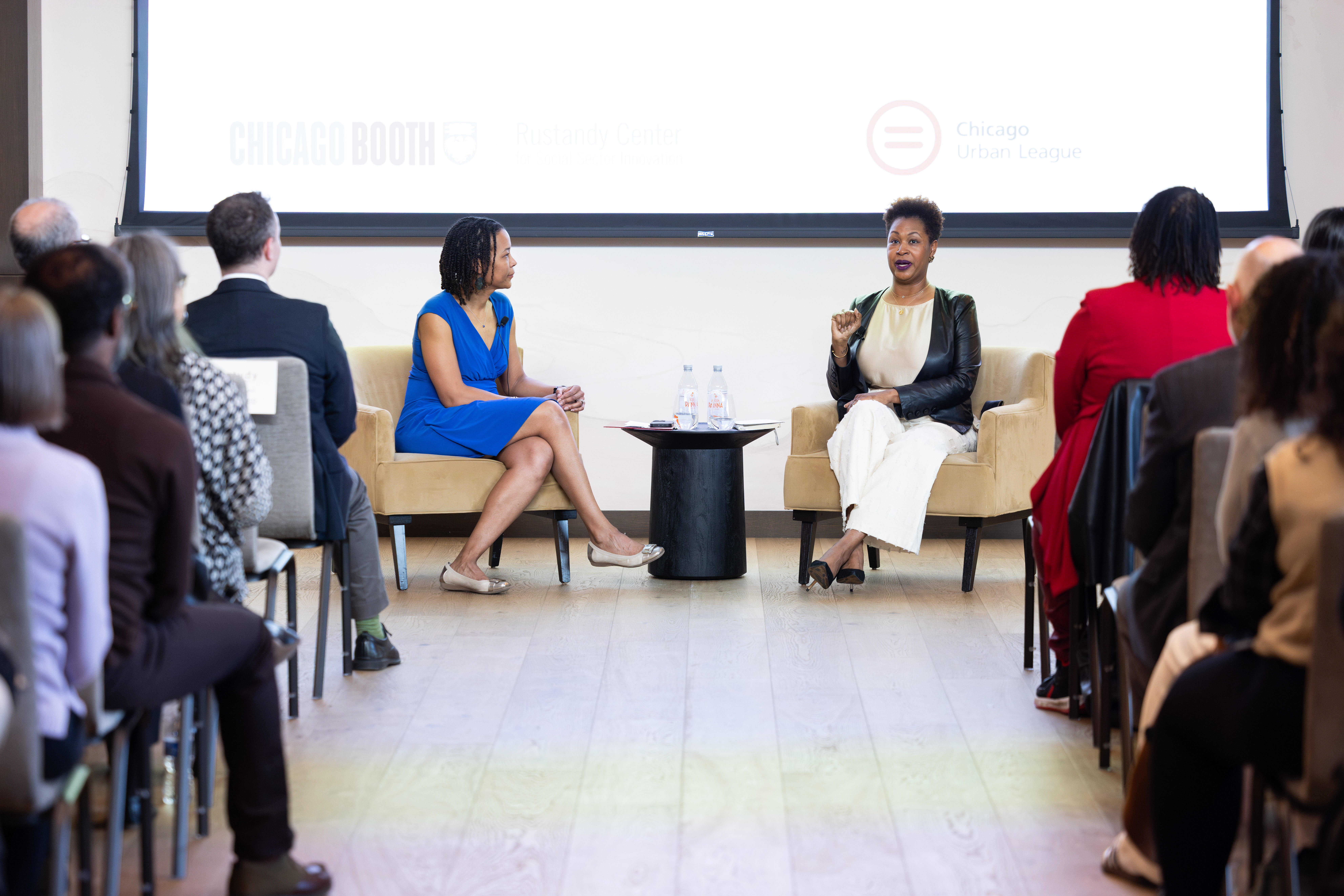Two women sitting in chairs conversing in front of people sitting in rows of chairs in a classroom or auditorium