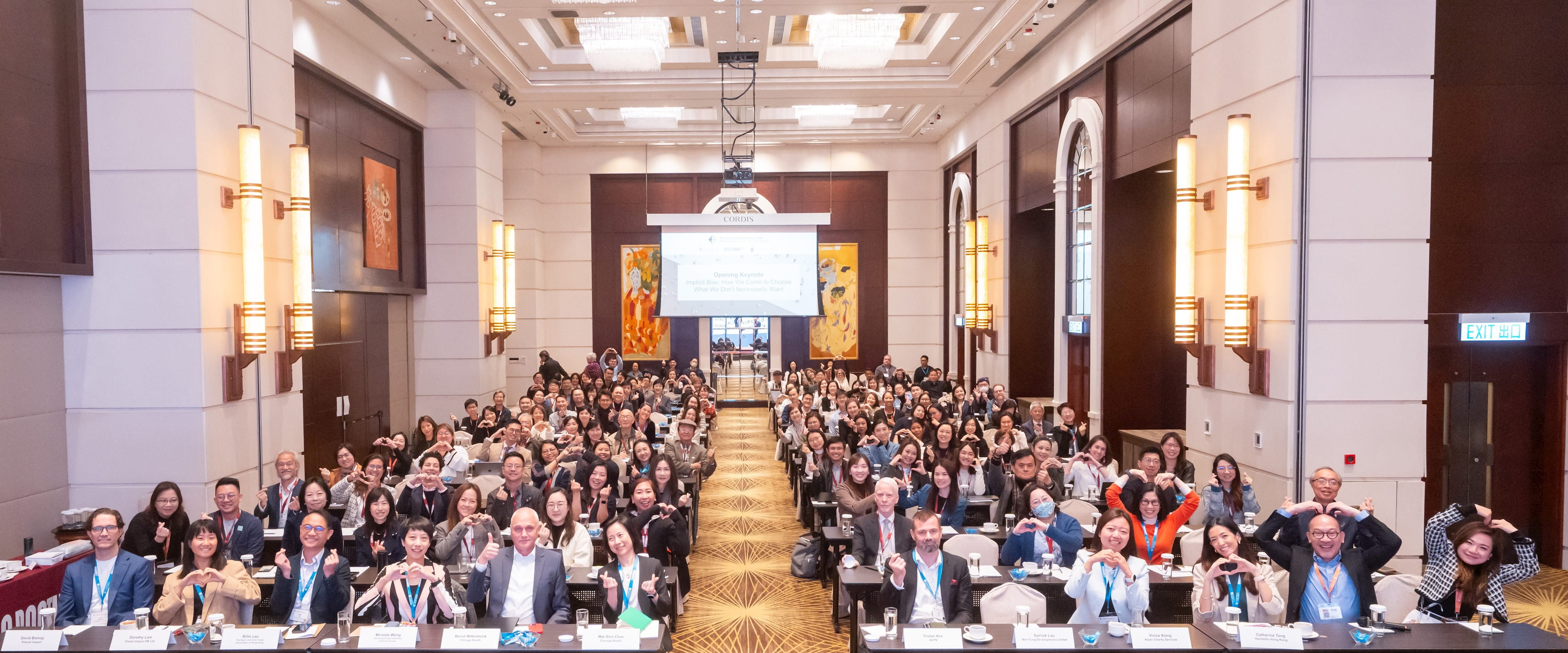 A large room with a screen at the back and many people sitting in rows at desks, making hand gestures