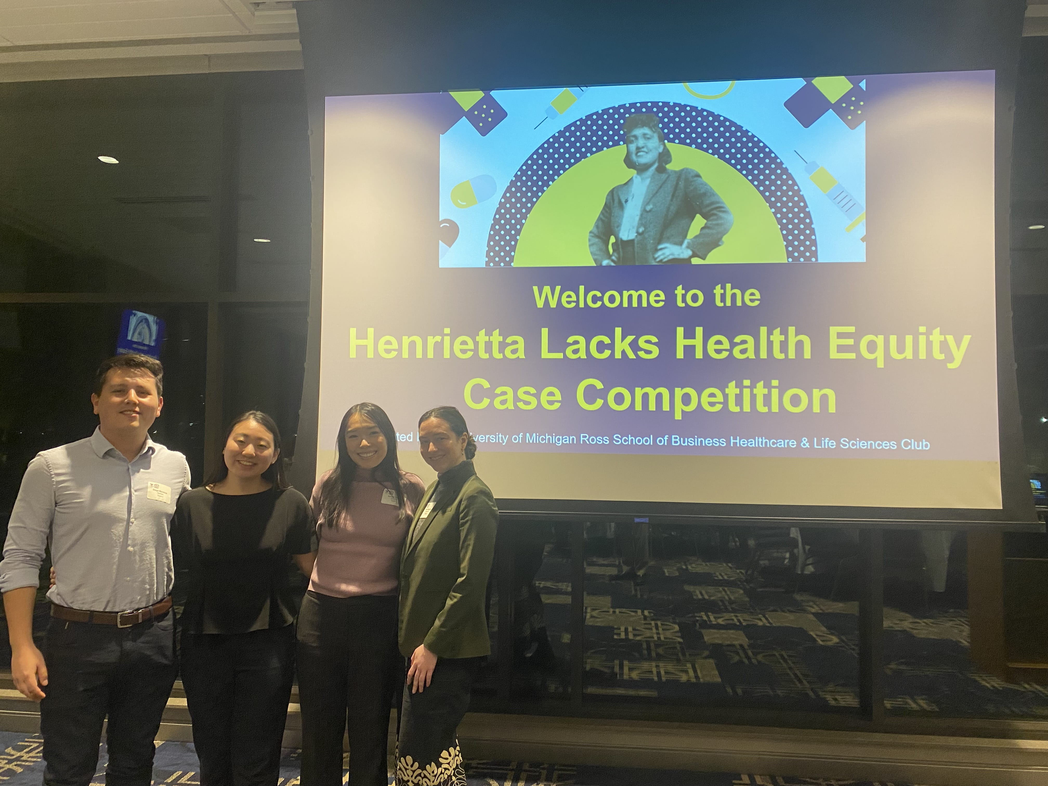 Four people standing in front of a large screen that reads Welcome to the Henrietta Lacks Health Equity Case Competition