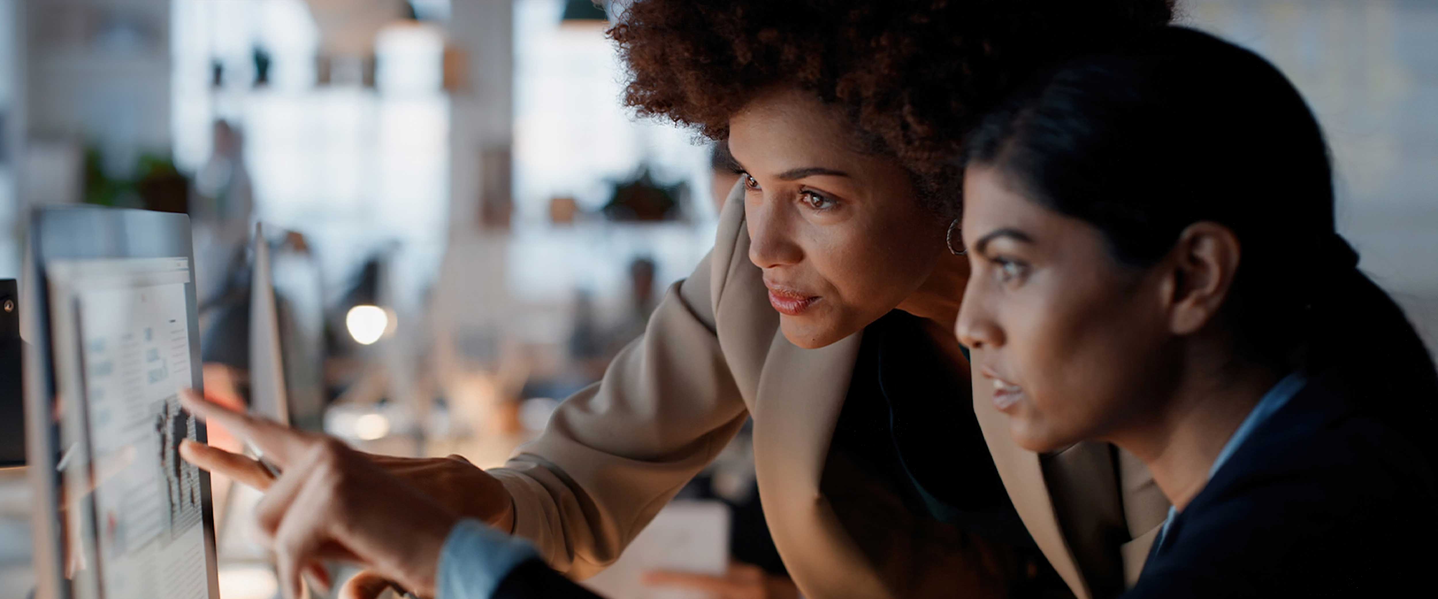 Two businesswomen work at a computer together, gesturing at screen