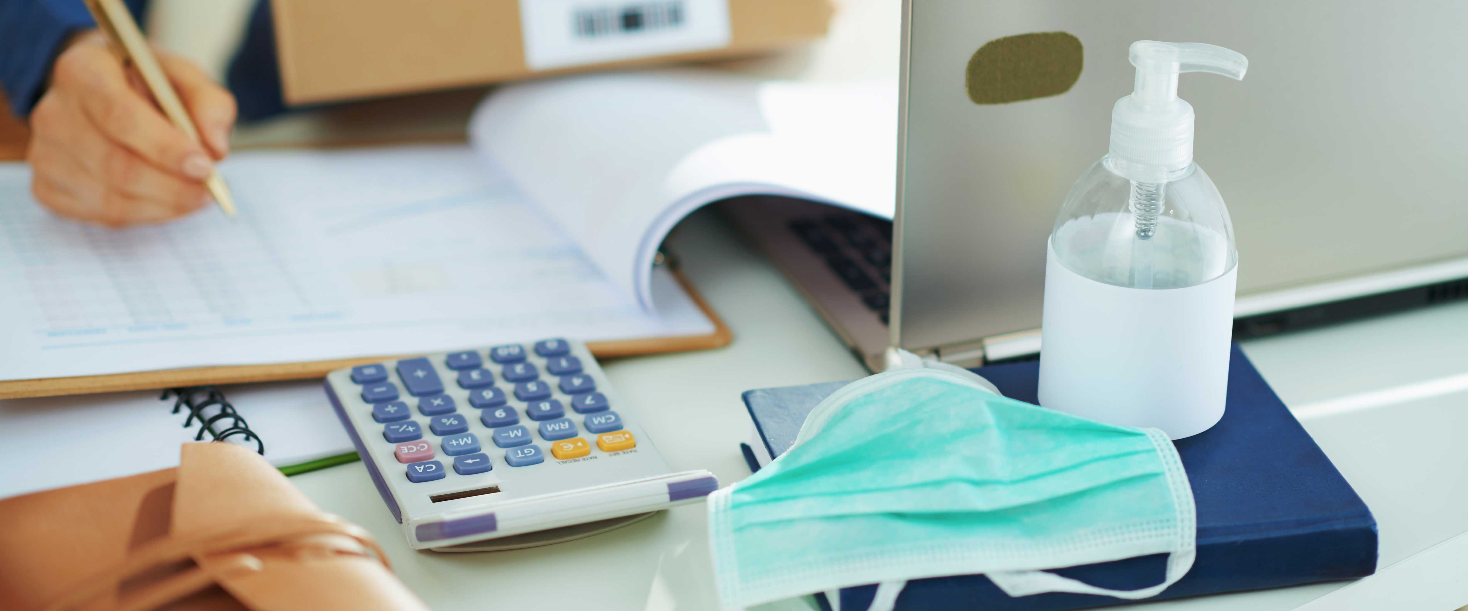 A hand writes at a desk with a clipboard, laptop, face mask and hand sanitizer
