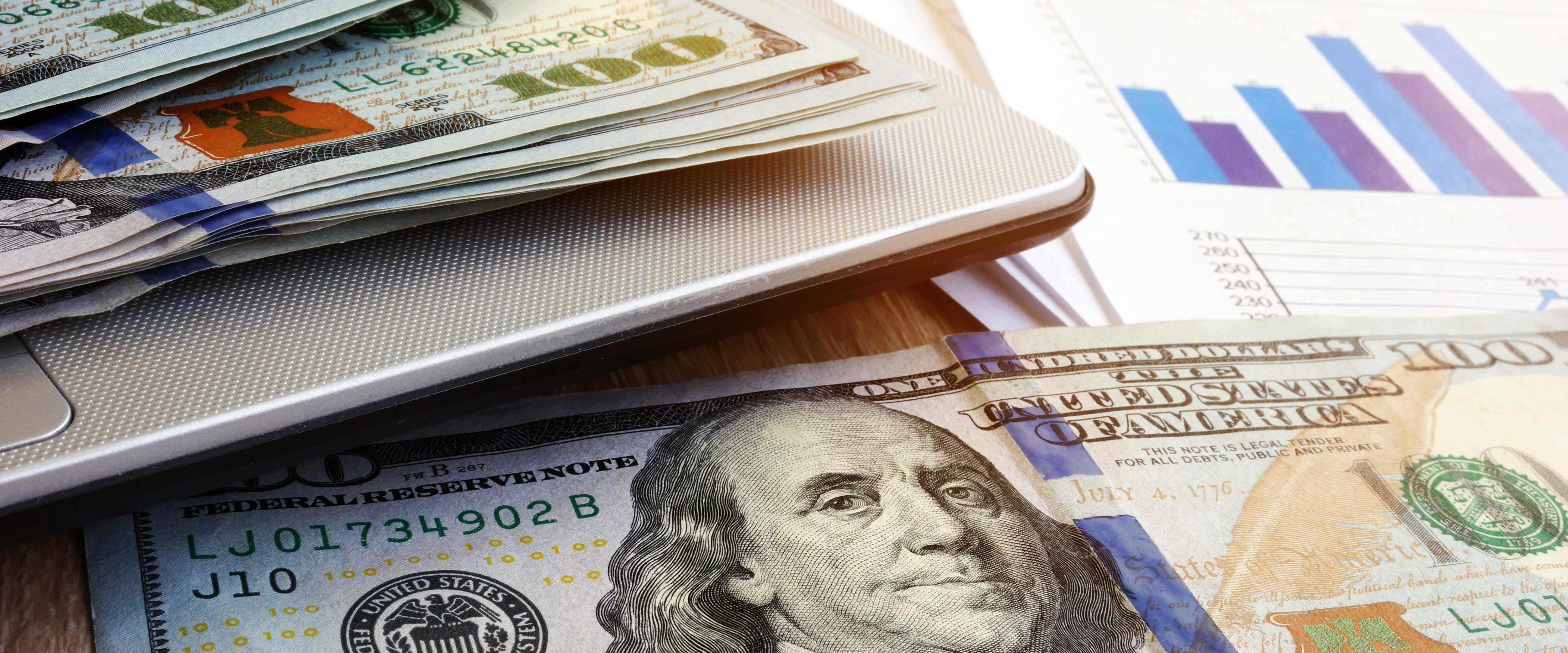 A laptop covered in hundred dollar bills and graph papers