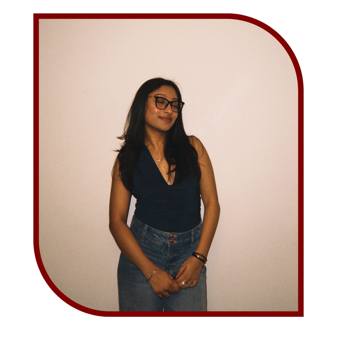 Picture of smiling dark-haired woman wearing a black blouse and jeans. She is closing her eyes in front of a cream background. The picture is bordered by a maroon frame. 