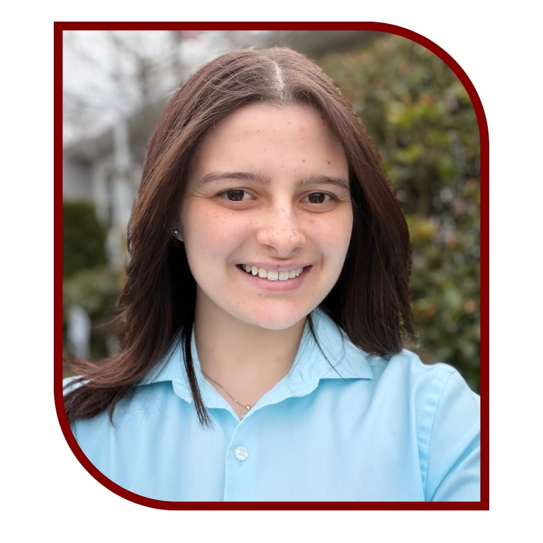 Picture of smiling dark-haired woman wearing a blue dress shirt and standing in front of a tree. The picture is bordered by a maroon frame. 