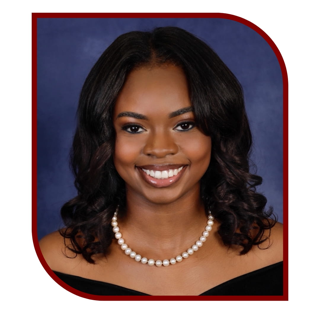 Picture of smiling dark-haired woman wearing a black dress and a pearl necklace standing in front of a blue background. The picture is bordered by a maroon frame. 