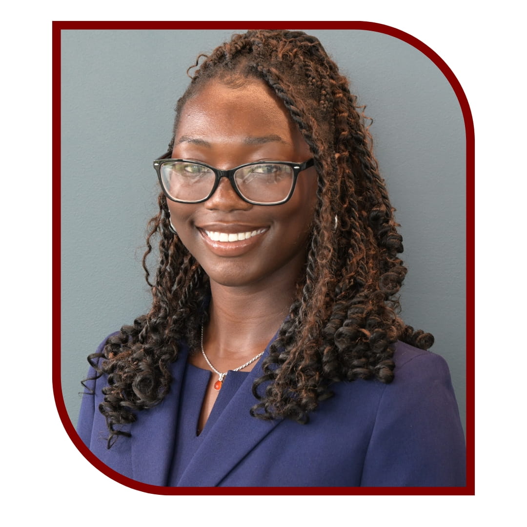 Picture of smiling woman with braids wearing a blue dress shirt and standing in front of a grey background. The picture is bordered by a maroon frame. 