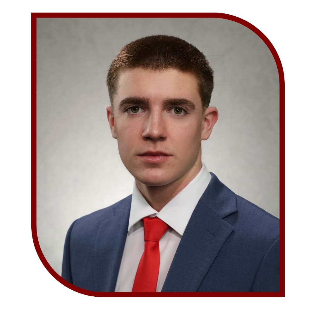 Picture of young man wearing a blue dress shirt and red tie and standing in front of grey background. The picture is bordered by a maroon frame. 