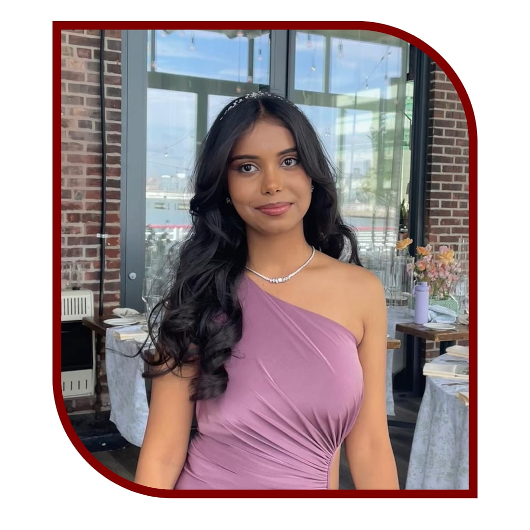 Picture of a dark-haired woman wearing a purple dress and standing in front of a restaraunt background. The picture is bordered by a maroon frame.