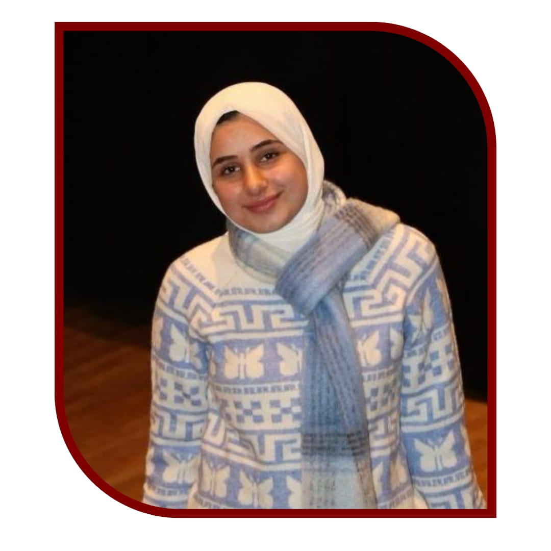 Picture of a smiling woman wearing a hijab and a blue sweater. She is standing in front of a stage background. The picture is bordered by a maroon frame.