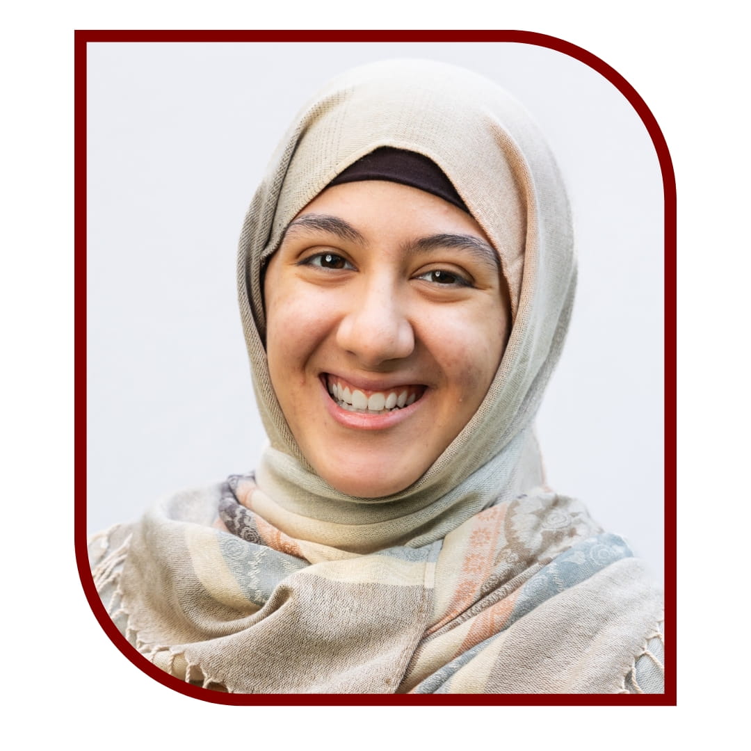 Picture of a smiling woman wearing a cream hijab and matching tunic. She is standing in front of a white background. The picture is bordered by a maroon frame.