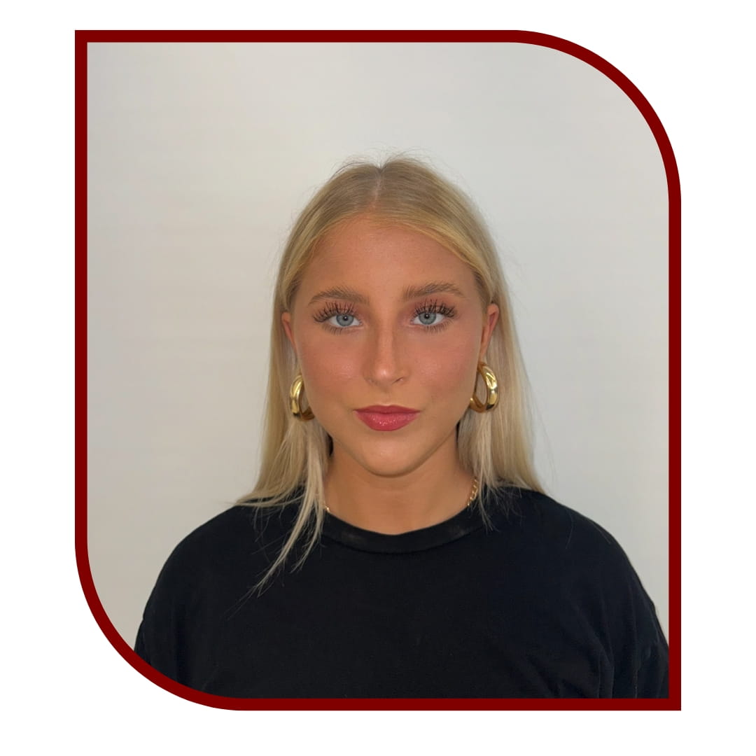 Picture of a blonde woman wearing a black blouse and standing in front of a white background. The picture is bordered by a maroon frame.