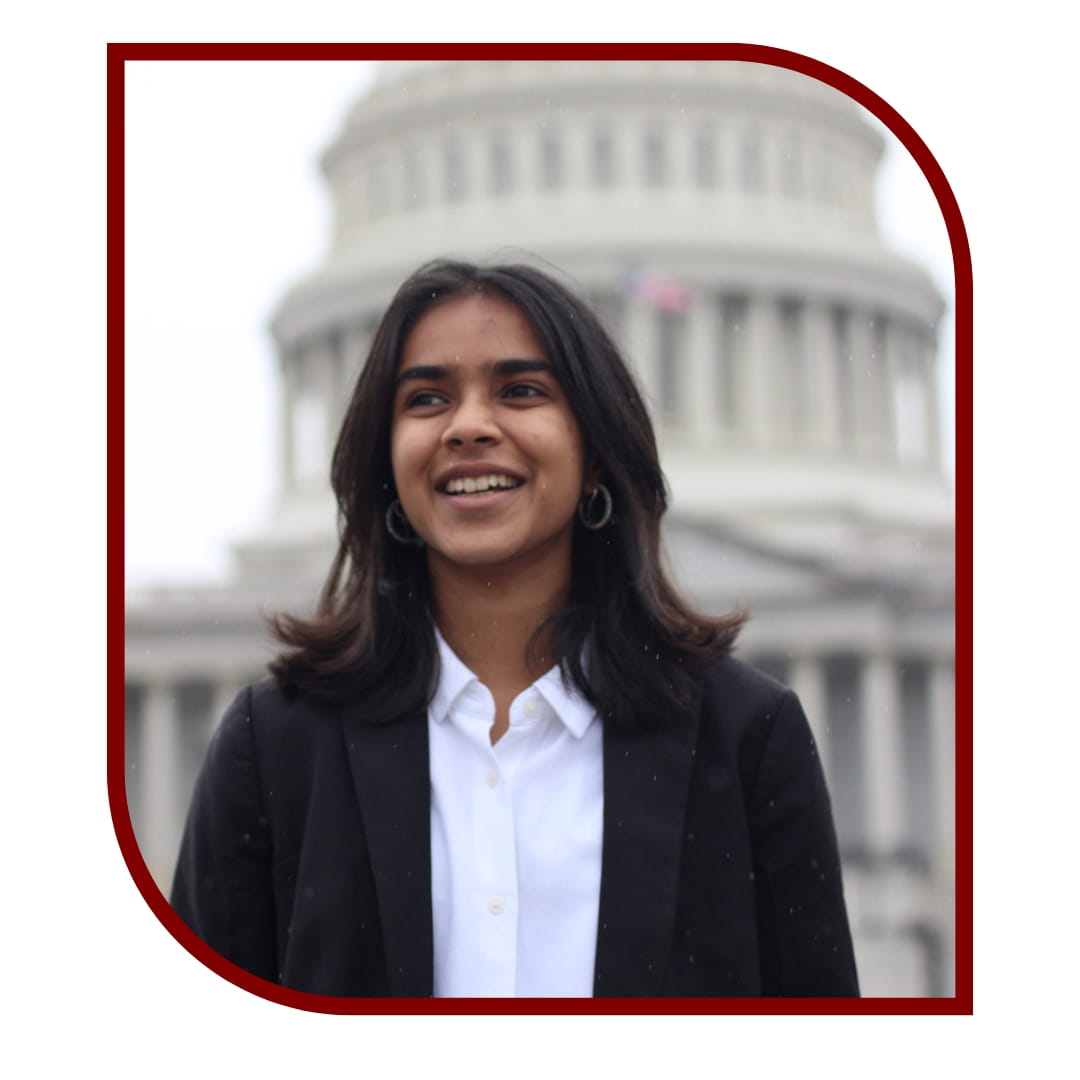 Picture of a smiling dark-haired woman wearing a black blazer and white undershirt and standing in front of the DC capitol. The picture is bordered by a maroon frame.
