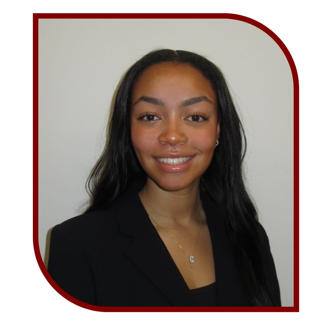 Picture of a smiling dark-haired woman wearing a black blazer and standing in front of a grey background. The picture is bordered by a maroon frame.