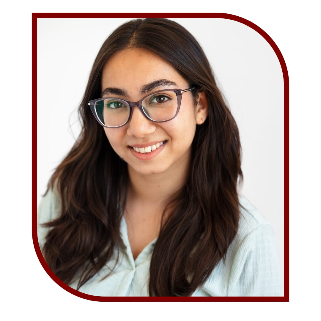 Picture of a smiling dark-haired woman wearing a white dress shirt and glasses and standing in front of a white background. The picture is bordered by a maroon frame.