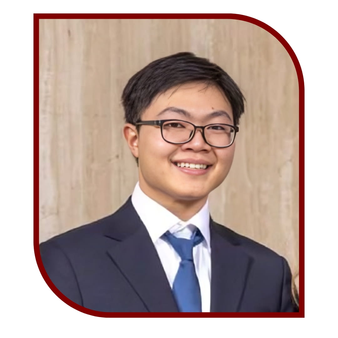 Picture of a smiling dark-haired man wearing a tuxedo and glasses and standing in front of a tan background. The picture is bordered by a maroon frame.