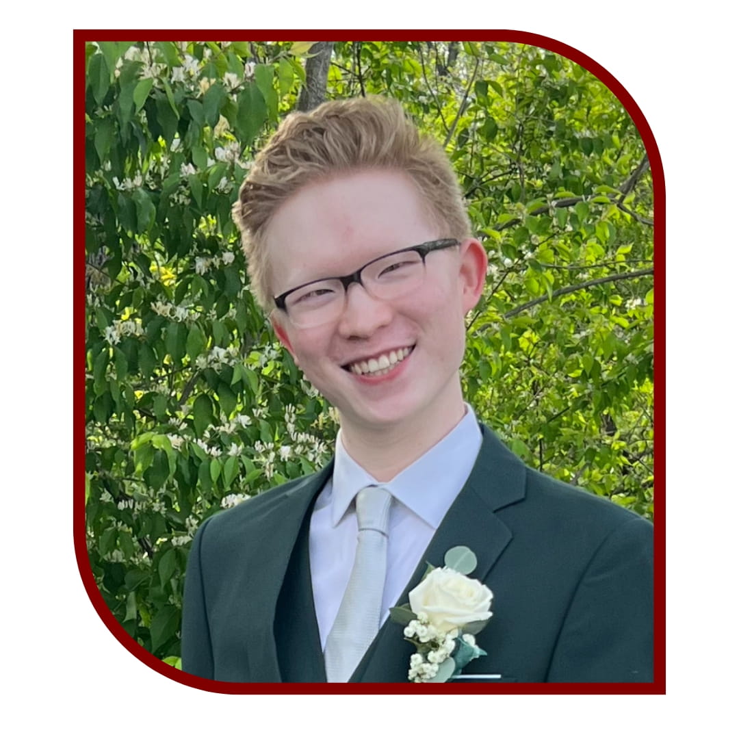 Picture of a smiling auburn-haired person wearing a tuxedo and glasses and standing in front of a leafy background. The picture is bordered by a maroon frame.