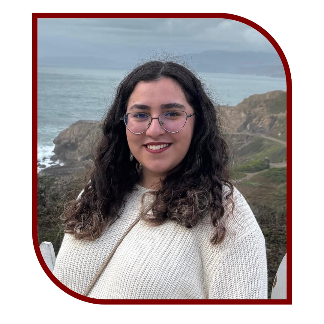 Picture of a smiling dark-haired woman wearing a white blouse and standing in front of a hill and sea. The picture is bordered by a maroon frame.