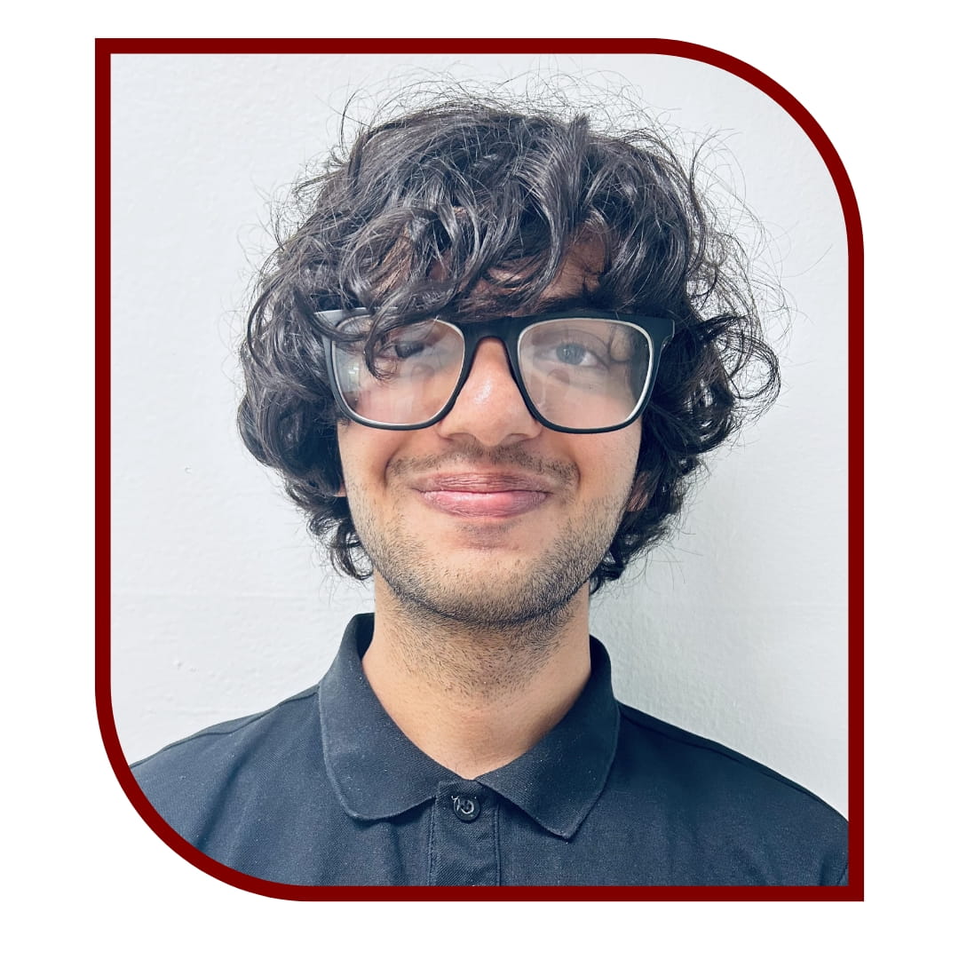 Picture of a smiling dark-haired man wearing a navy shirt and glasses and standing in front of white background. The picture is bordered by a maroon frame.