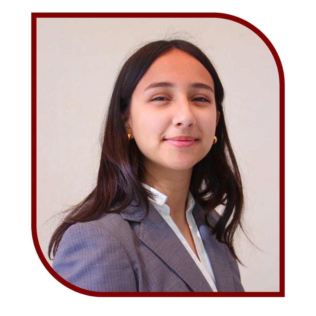 Picture of a smiling dark-haired woman wearing a blazer and standing in front of a white background. The picture is bordered by a maroon frame.