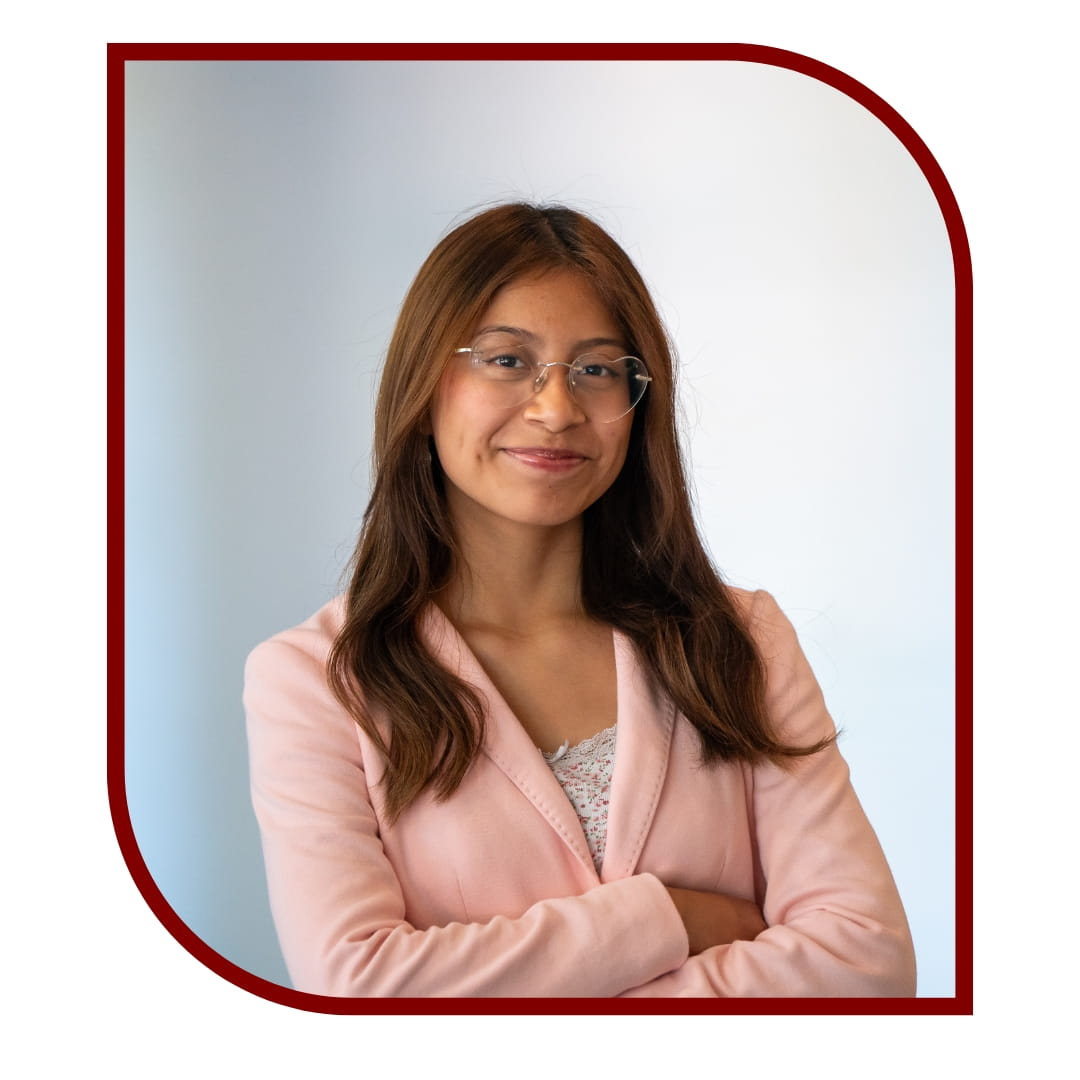 Picture of a smiling brown-haired woman wearing a pink blazer and standing in front of a white background. The picture is bordered by a maroon frame.