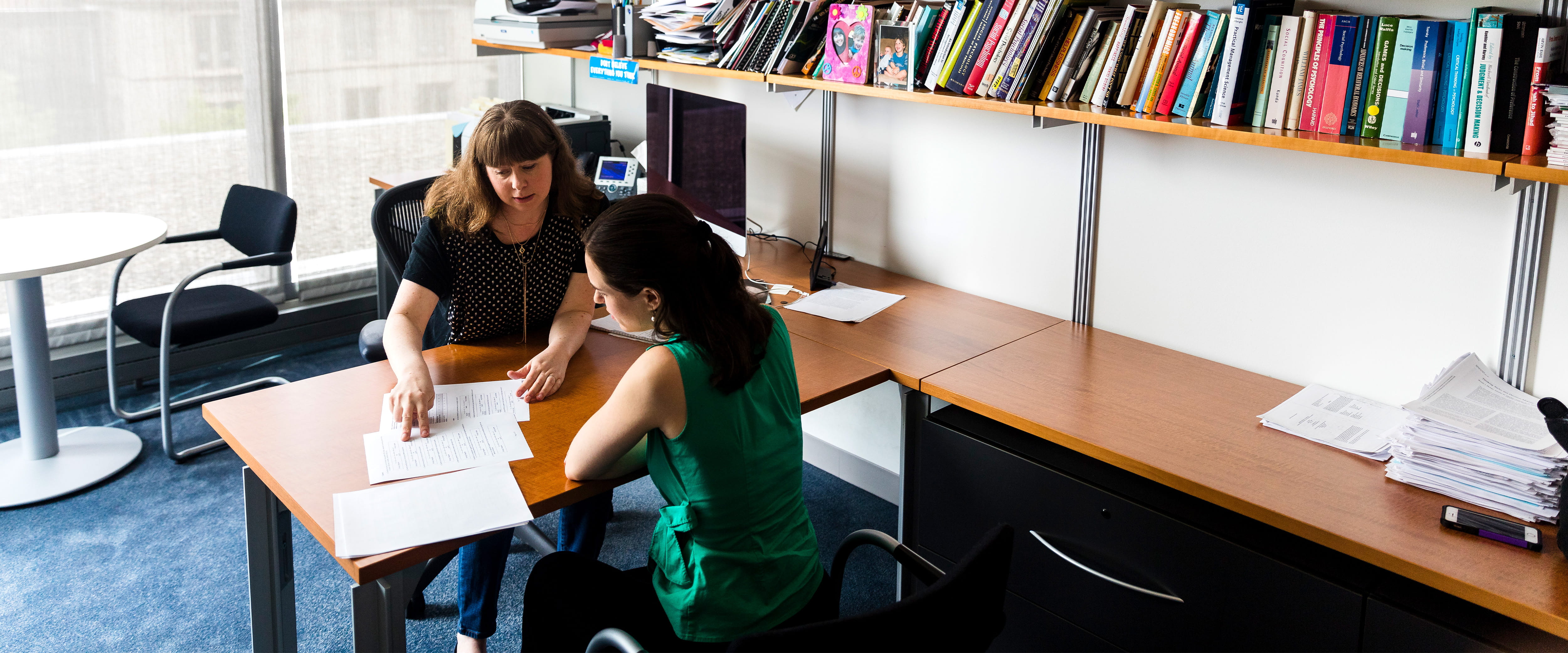 A woman points to a piece of paper at her desk while a young student looks it over.
