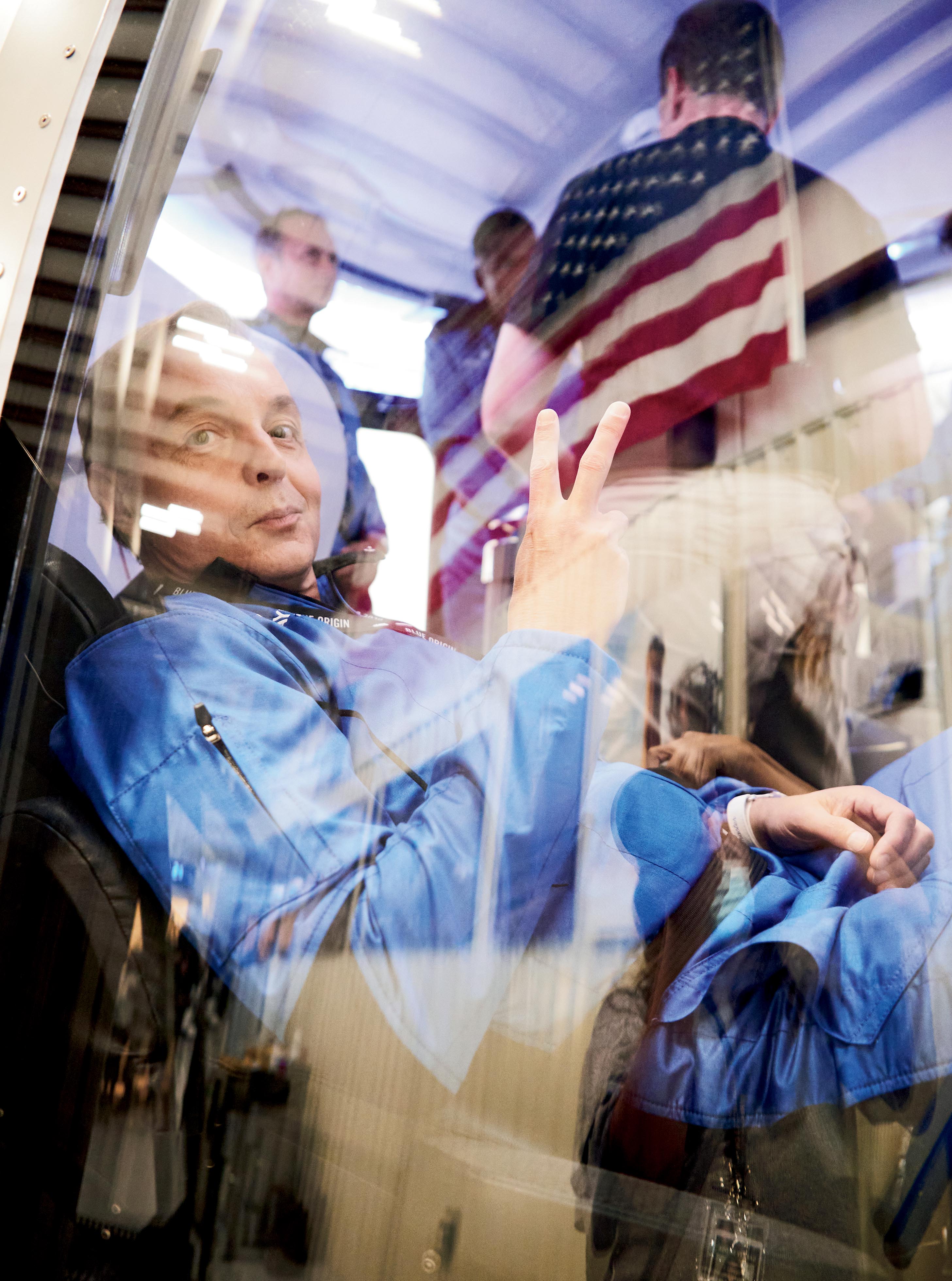 A crew member of Blue Origin's space exploration program giving a peace sign through a window