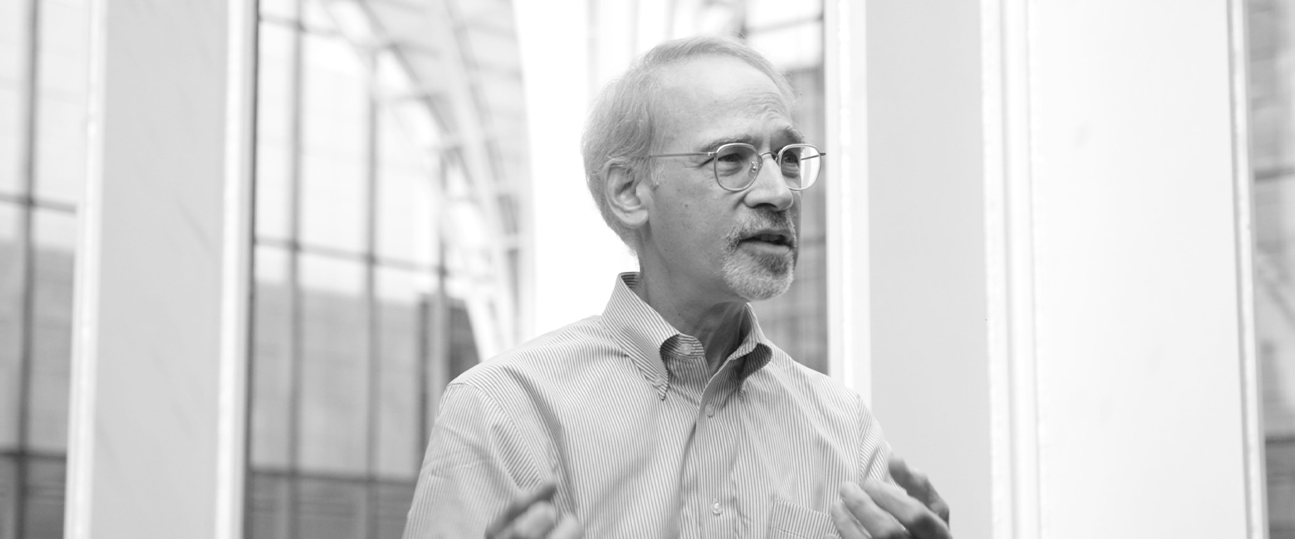 Black and white photo of James Schrager in front of a wall of windows, gesturing while talking.