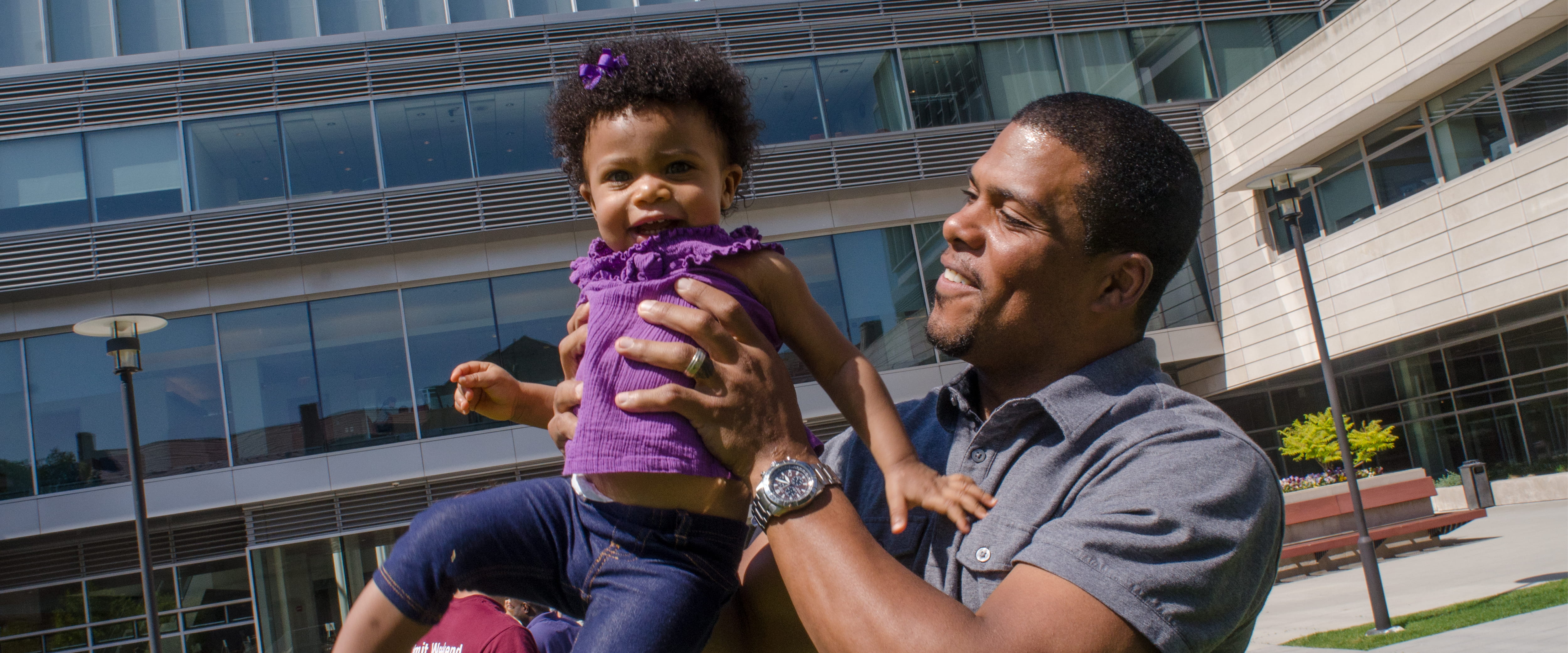 A smiling male student holds his daughter, outside Harper Center