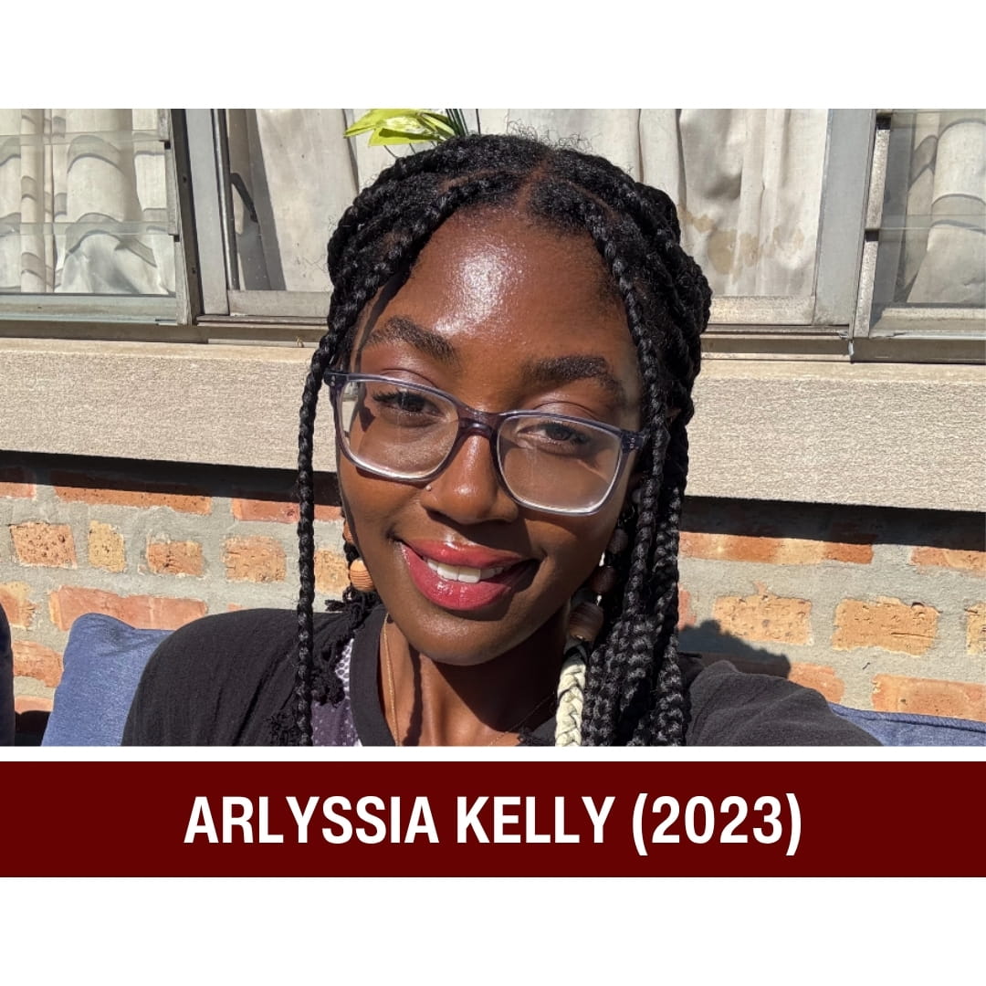 Picture of smiling dark-haired woman wearing a white and black shirt and standing in front of a window. A red strip beneath the picture is overlaid with white text that reads: Arlyssia Kelly (2023)